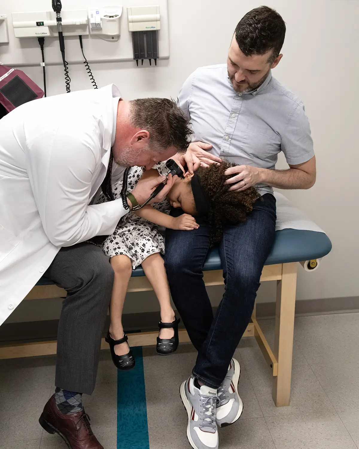 Modern day photo of family physician Dr. Peter Bidey examining a young child on a father's lap