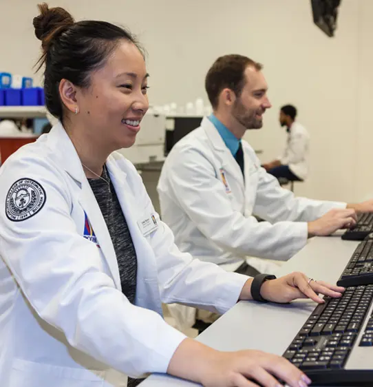 Pharmacy students at PCOM Georgia smile as they work on computers in a lab in their white coats.