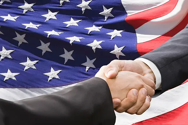 Two people shaking hands with an American flag in the background.