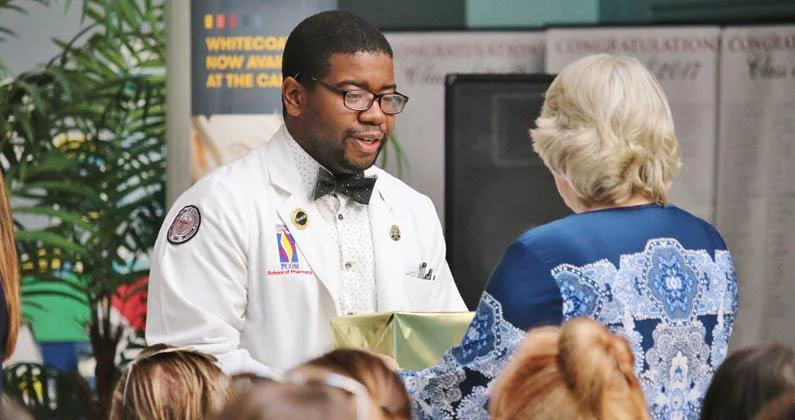 A PCOM Georgia student, wearing a white coat and black bow tie, hands a box of cremains to a guest at the memorial service.