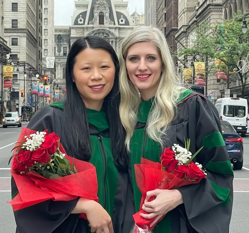 Julia Burns and Amanda Bond stand side by side while wearing commencement robes and holding bouquets