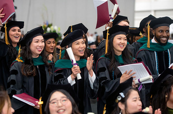 PCOM graduates dressed in regalia cheer and clap during commencement ceremony