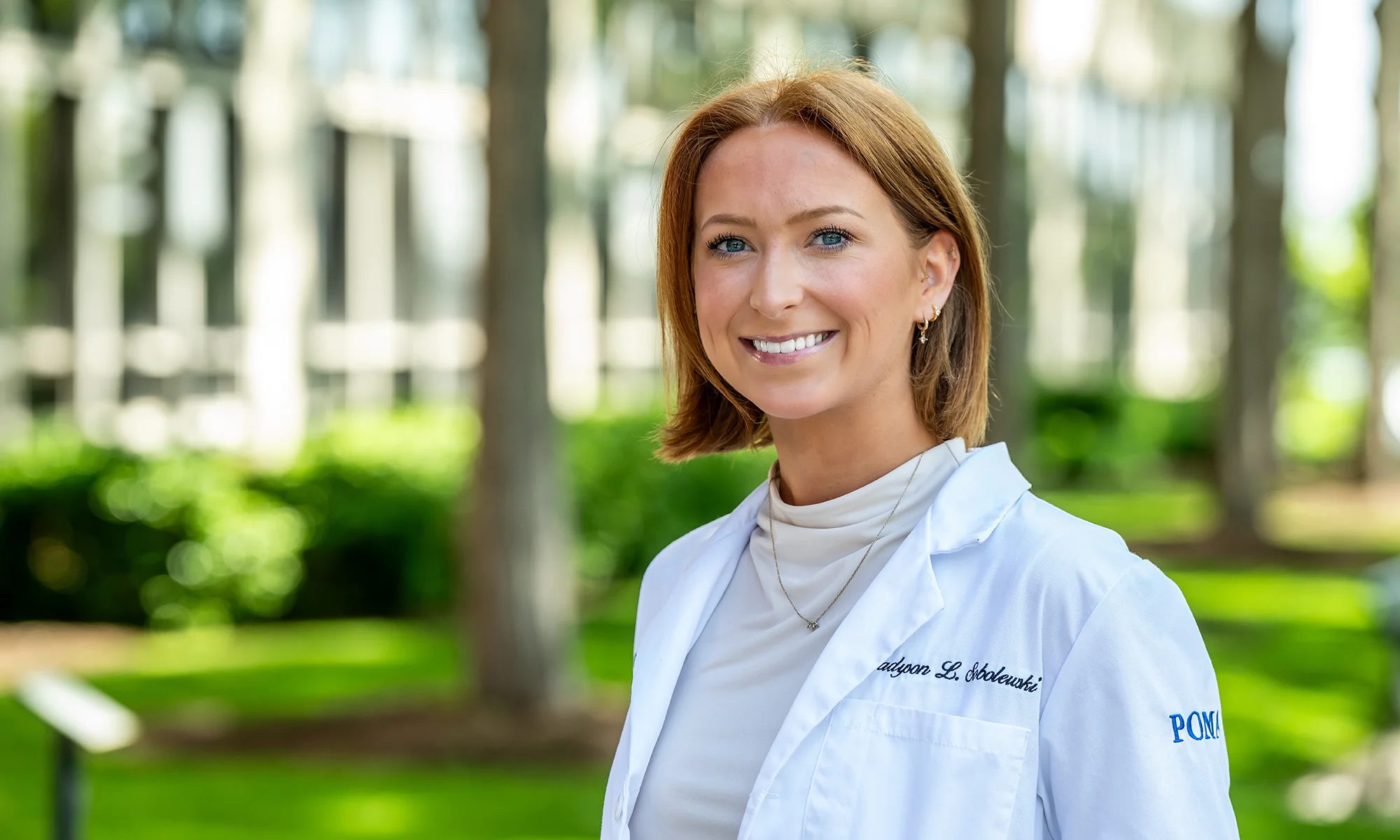 A PCOM medical student smiles outside while wearing the student physician white coat