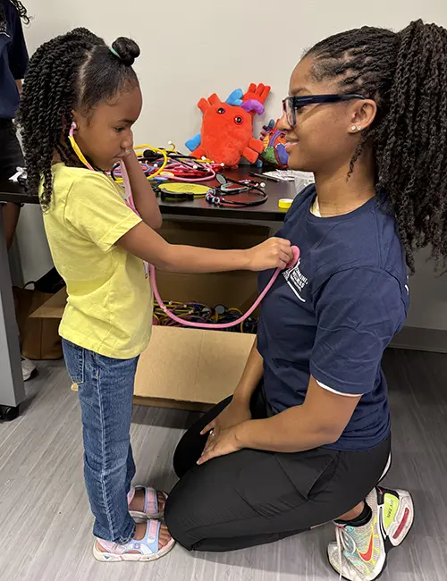 A young child uses a stethoscope to listen to the heart of a PCOM medical student at the Lancaster Ave Healthcare Center community day celebration