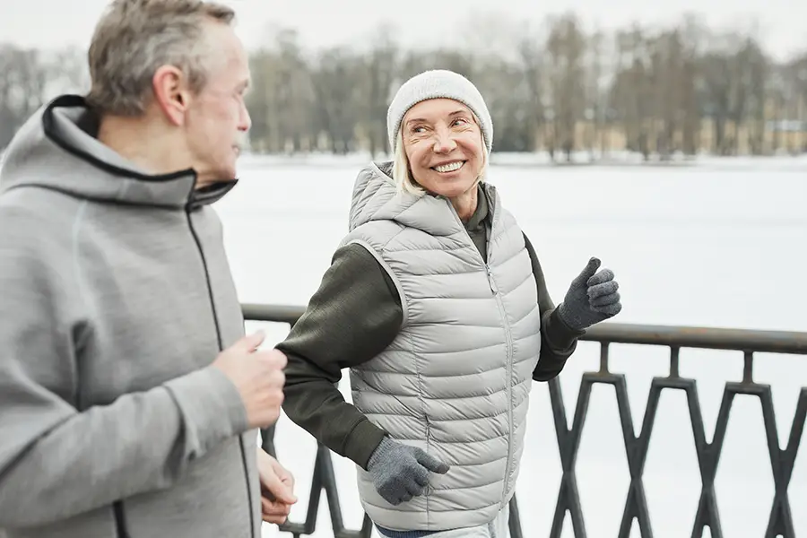 A senior couple smiles as they get exercise by walking through a snowy town in order to maintain their health during the winter season