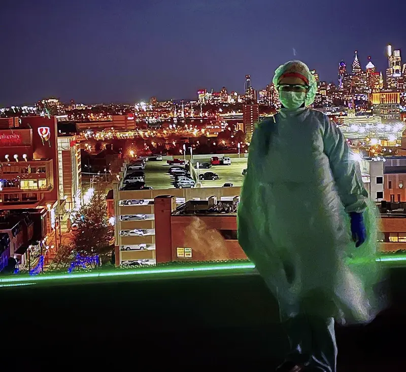 PCOM surgery resident Christina Monaco Poloni stands in her surgery scrubs on a rooftop with the Philadelphia skyline in the background.