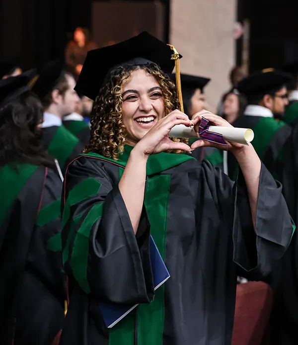 A PCOM South Georgia DO graduate smiles and makes a heart symbol with her hands while holding her diploma