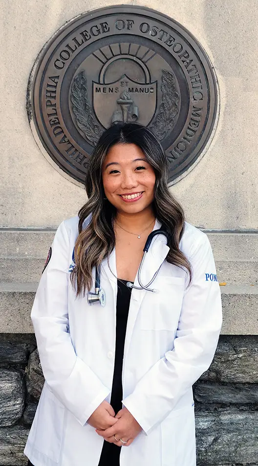 PCOM osteopathic medical student  Alexandra Wong smiles with her white coat in front of the College seal and obelisk