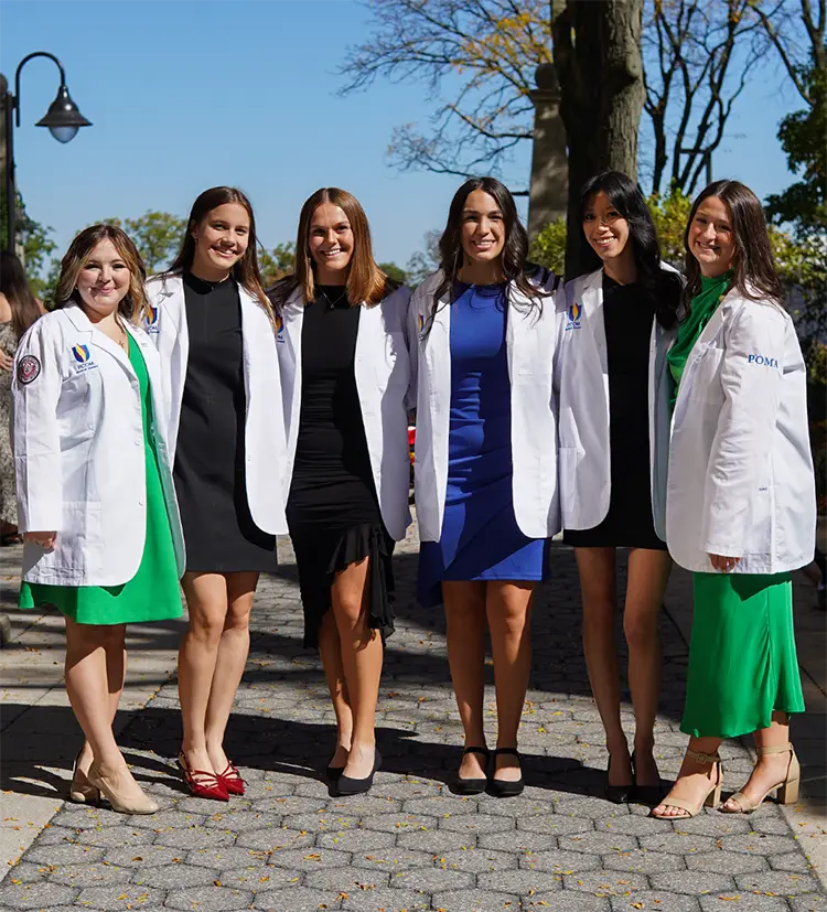 Six medical students smile donning their white coats outside on a pathway at PCOM's Philadelphia campus