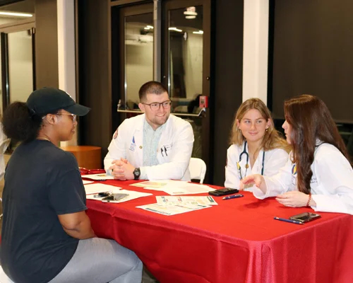 Three DO students in white lab coats sit at a table with a women wearing a black hat and a black shirt.