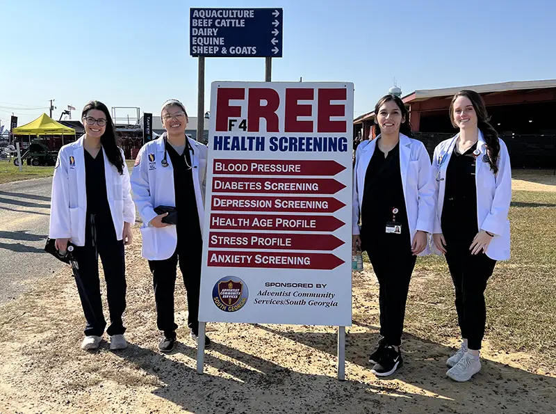 PCOM DO students smile in their white coats and hold their stethoscopes in front of a Free Health Screening sign at the Sunbelt Ag Expo