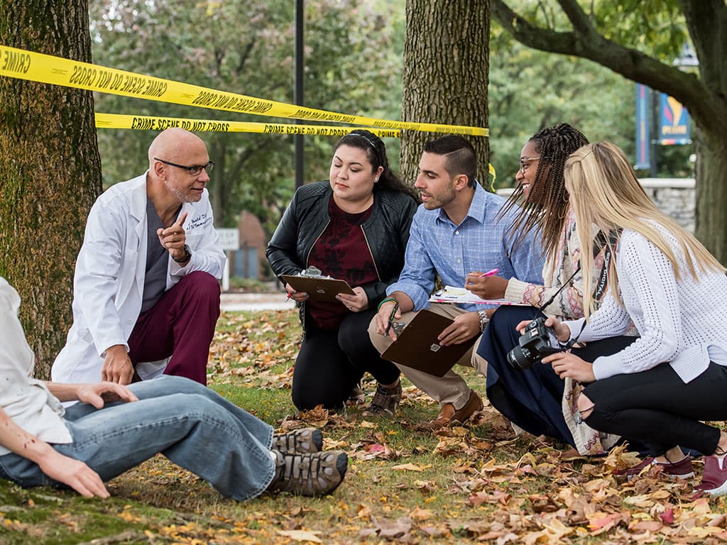 Forensic medicine students crouch at mock crime scene with professor Gregory McDonald