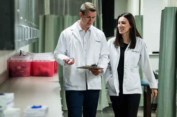 Two physician assistant students smile as they walk and talk over an exam chart in a lab at PCOM