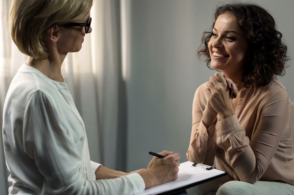 A mental health counselor and patient smile as they interact on a couple of couches in an office.